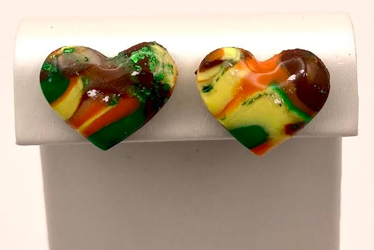 A pair of heart-shaped stud earrings with a marbled, swirled pattern of green, orange, yellow, brown, and white, displayed on a white rectangular stand.