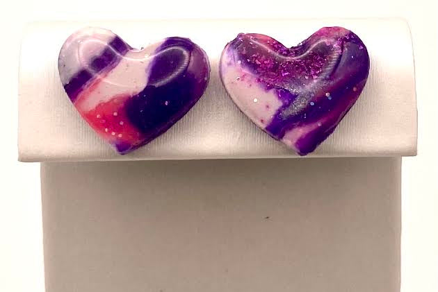 A pair of heart-shaped glass stud earrings with a purple, pink, and white galaxy swirl design, displayed on a white jewelry stand.