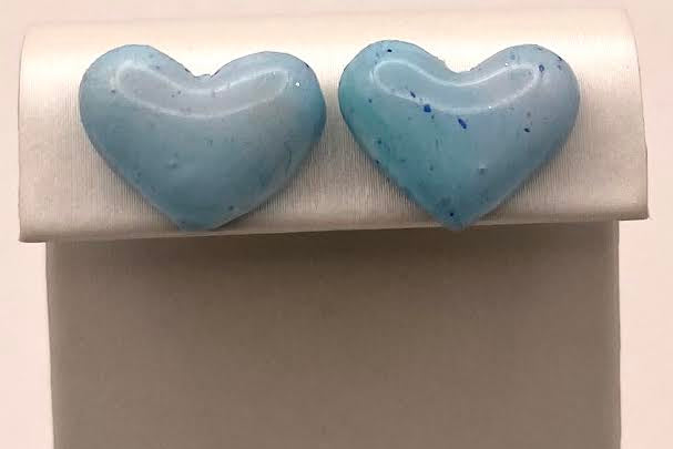 A pair of light blue heart-shaped stud earrings displayed on a white jewelry stand against a plain light background.