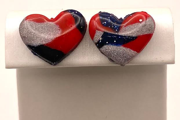 A pair of heart-shaped glass stud earrings with a red, white, and blue marbled pattern, displayed on a white jewelry stand.