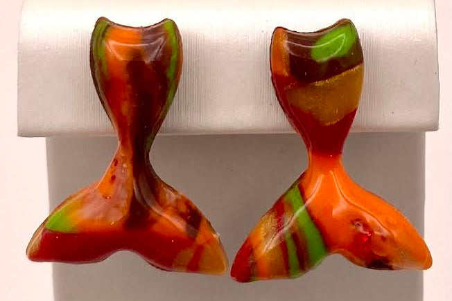 A pair of handmade glass stud earrings shaped like mermaid tails, featuring a marbled pattern of orange, red, green, and brown, displayed on a white jewelry stand.