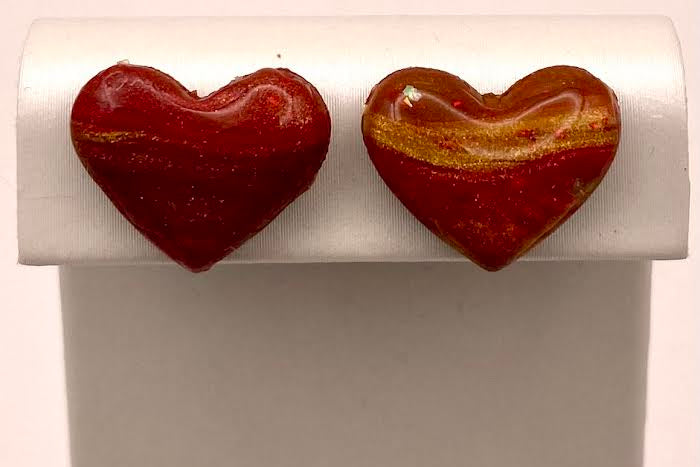 A pair of heart-shaped glass stud earrings with a marbled red and gold pattern, displayed on a white rectangular jewelry stand.