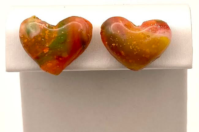 A pair of heart-shaped glass stud earrings with a marbled pattern of orange, red, yellow, and green, displayed on a white jewelry stand.