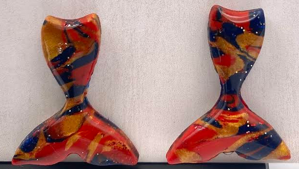 A pair of colorful, abstract mermaid tail earrings displayed on a black stand in front of a light beige textured card with the brand name 'Phoenix Frost Art' printed on it.