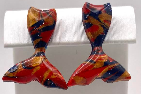 A pair of marbled resin earrings shaped like mermaid tails, featuring swirls of red, orange, and dark blue, displayed on a white jewelry stand.