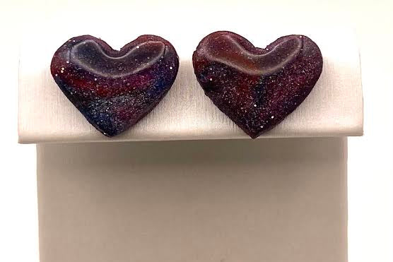 A pair of heart-shaped glass stud earrings with a marbled purple, red, and black pattern, displayed on a white rectangular jewelry stand against a white background.