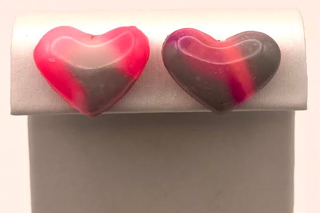 A pair of heart-shaped stud earrings with a marbled pink, red, and dark gray pattern, displayed on a gray jewelry stand against a white background.