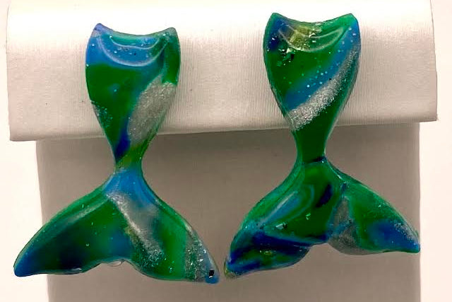 A pair of handmade mermaid tail stud earrings with a swirled green, blue, and white glass design, displayed on a white jewelry stand.