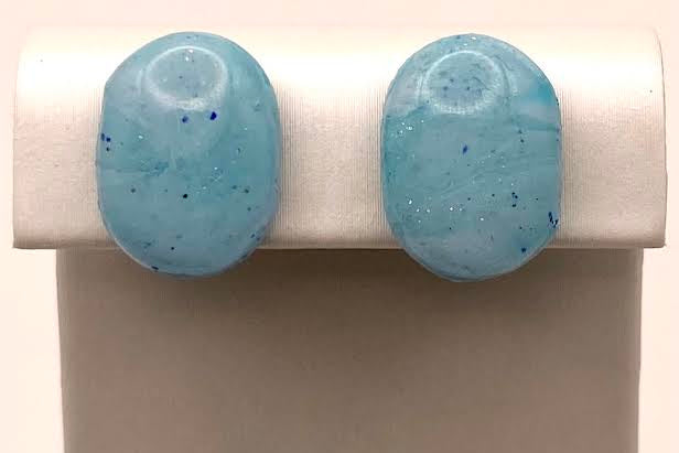 A pair of oval stud earrings in a light blue marbled glass finish with tiny dark specks, displayed on a white jewelry stand against a light background.