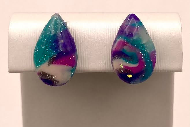 A pair of teardrop-shaped stud earrings with a marbled swirl pattern of purple, teal, white, and pink, accented with glitter, displayed on a white jewelry stand.