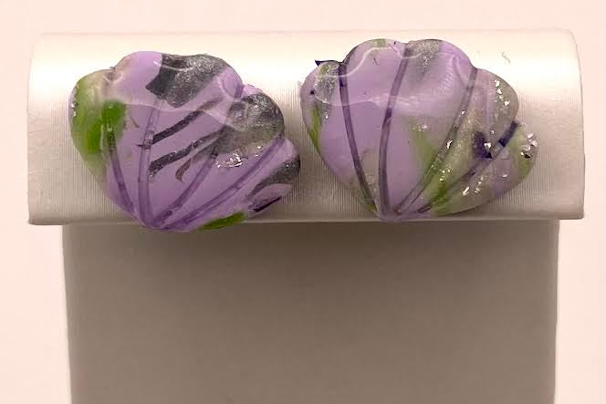 A pair of handmade lavender and purple seashell-shaped earrings with green and black marbled details, displayed on a white jewelry stand.