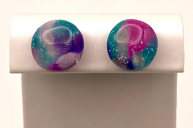 A pair of round stud earrings with a marbled swirl pattern of purple, teal blue, and white, displayed on a white plastic jewelry stand against a white background.