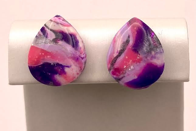 A pair of teardrop-shaped stud earrings with a marbled swirl pattern of purple, pink, and white, displayed on a white jewelry stand against a light pink background.