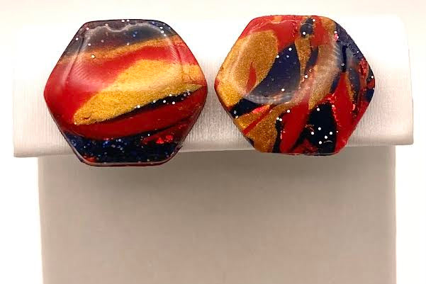 A pair of hexagonal stud earrings with a marbled swirl pattern of red, orange, yellow, and dark blue, displayed on a white plastic jewelry stand against a white background.