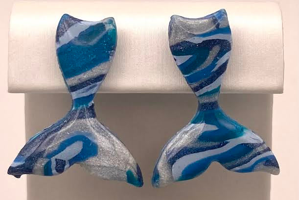 A pair of handmade mermaid tail stud earrings with a blue, white, and gray marbled pattern, displayed on a white rectangular stand.