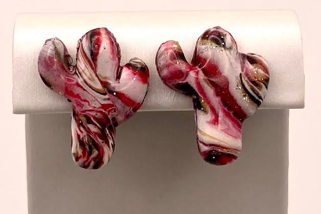 A pair of cactus-shaped stud earrings with a marbled pattern of red, white, and black, displayed on a white stand against a light background.