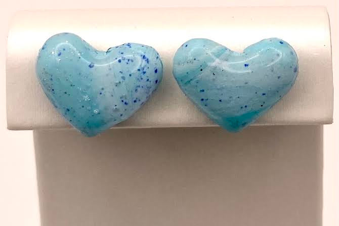 A pair of heart-shaped stud earrings in a light blue translucent resin with dark blue speckles, displayed on a white jewelry stand.