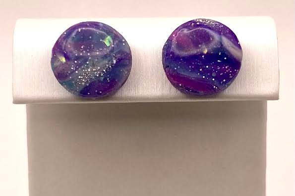 A pair of round stud earrings with a purple and blue galaxy design and glitter, displayed on a white jewelry stand.