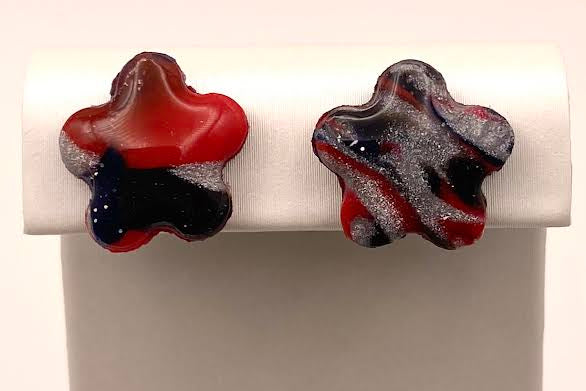 Two star-shaped stud earrings displayed on a white jewelry stand. One earring has a red, white, and dark blue marbled pattern, and the other has a red, white, and black marbled pattern.