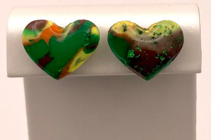 A pair of heart-shaped stud earrings with a marbled green, yellow, orange, and dark red pattern, displayed on a white jewelry stand against a light background.
