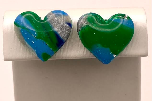 A pair of heart-shaped glass stud earrings with a marbled pattern of green, blue, and silver, displayed on a white jewelry stand.