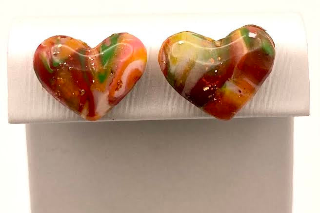 A pair of heart-shaped stud earrings with a marbled swirl pattern of orange, red, green, and white, displayed on a white jewelry stand.