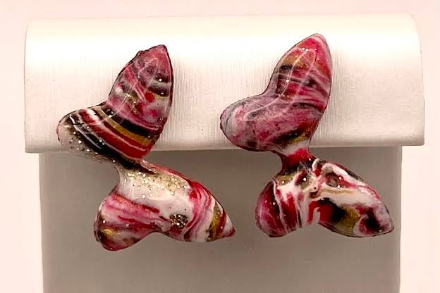 A pair of butterfly-shaped stud earrings with a marbled pattern of pink, red, white, and black, displayed on a white jewelry stand.