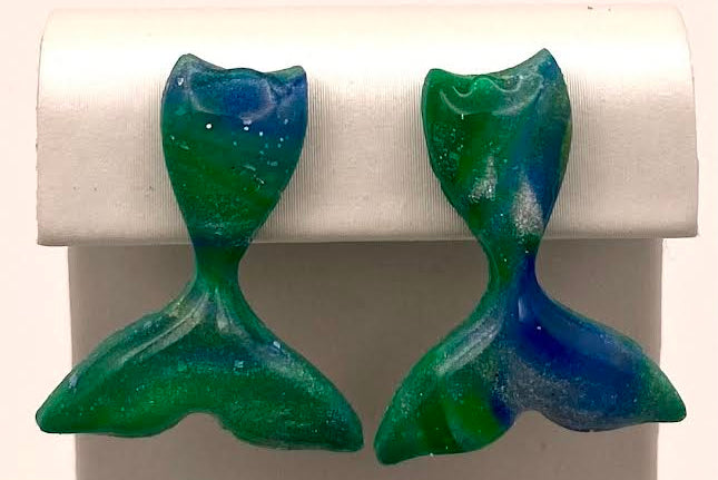 A pair of handmade mermaid tail stud earrings in a green and blue marbled glass finish, displayed on a white jewelry stand.
