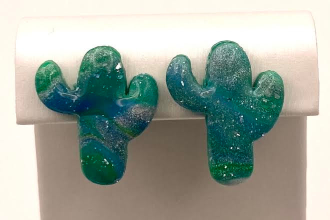 A pair of cactus-shaped stud earrings with a marbled blue and green glittery finish, displayed on a white jewelry stand against a white background.