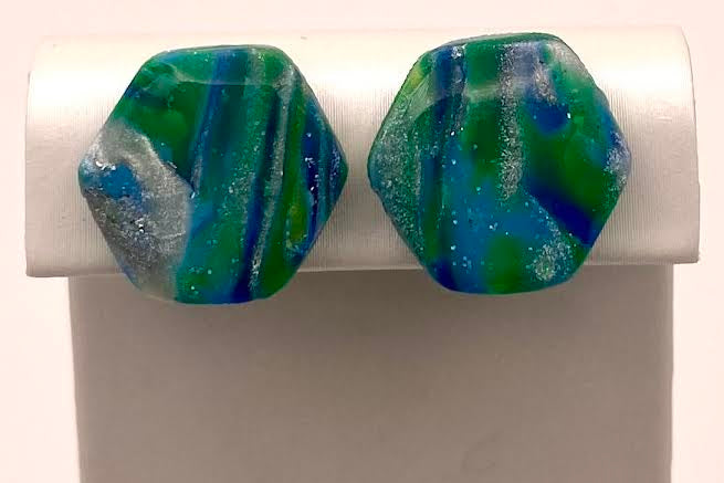 A pair of hexagonal stud earrings with a marbled green, blue, and white pattern, displayed on a white jewelry stand against a light background.