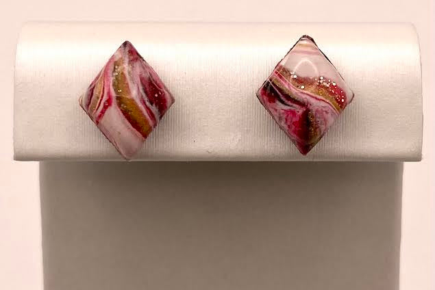 A pair of diamond-shaped stud earrings with a marbled pattern of pink, white, and gold, displayed on a white jewelry stand against a light beige background.