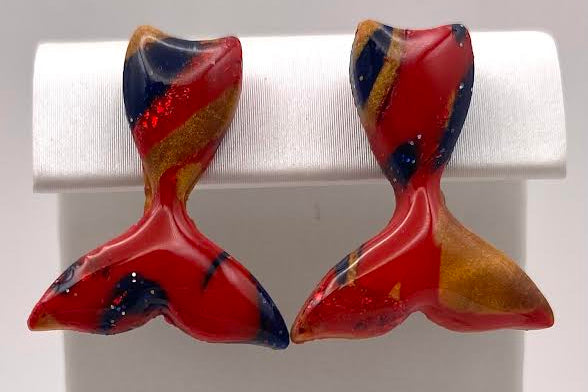 A pair of handmade fish-shaped earrings with a glossy red, orange, and navy blue marbled pattern, displayed on a white jewelry stand.
