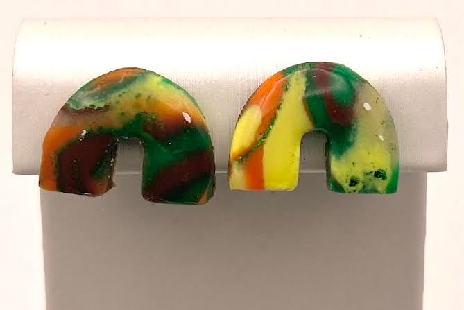A pair of handmade resin earrings with a marbled pattern of green, yellow, orange, and dark brown, displayed on a white plastic stand.