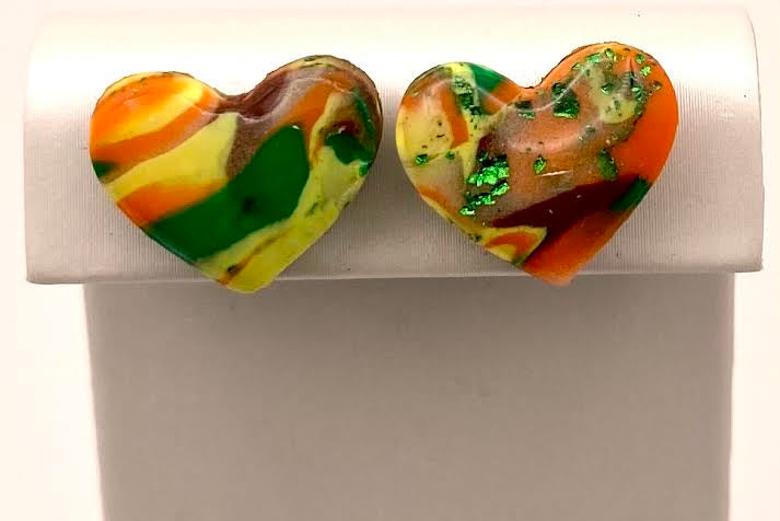 A pair of heart-shaped stud earrings with a marbled pattern of orange, green, yellow, and brown, displayed on a white jewelry stand against a white background.