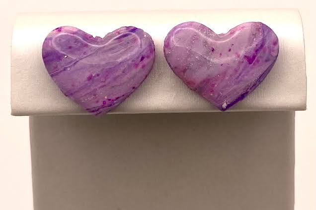 A pair of heart-shaped stud earrings with a marbled purple and white pattern, displayed on a white jewelry stand against a light background.