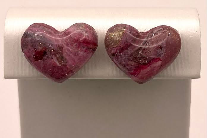 A pair of heart-shaped stud earrings with a marbled pink, red, and white pattern and small gold flecks, displayed on a white jewelry stand.