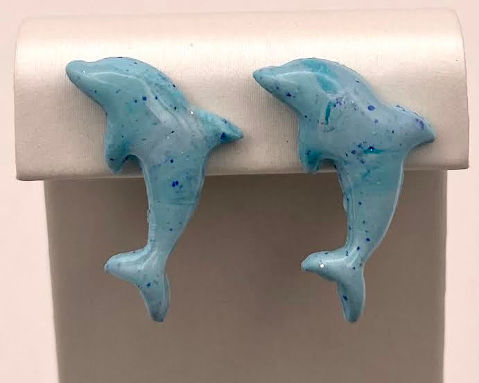 A pair of light blue dolphin-shaped stud earrings with darker blue speckles, displayed on a white jewelry stand against a light background.
