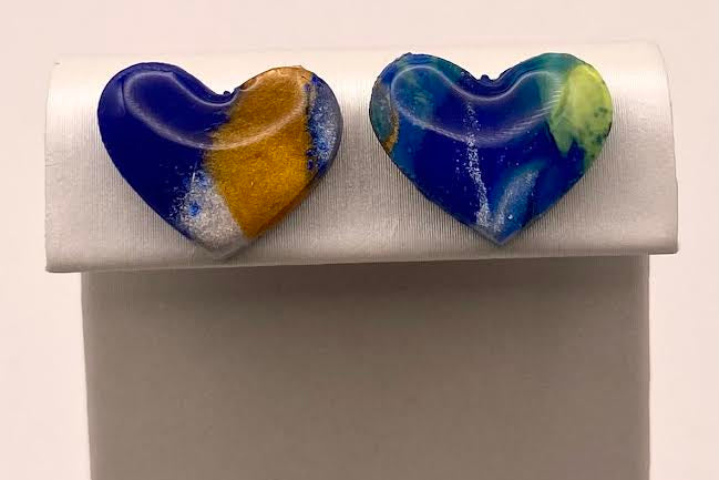 A pair of heart-shaped glass stud earrings displayed on a white jewelry stand. One earring is split vertically into blue and yellow halves with white marbling, and the other features swirling blue, white, and light green patterns.
