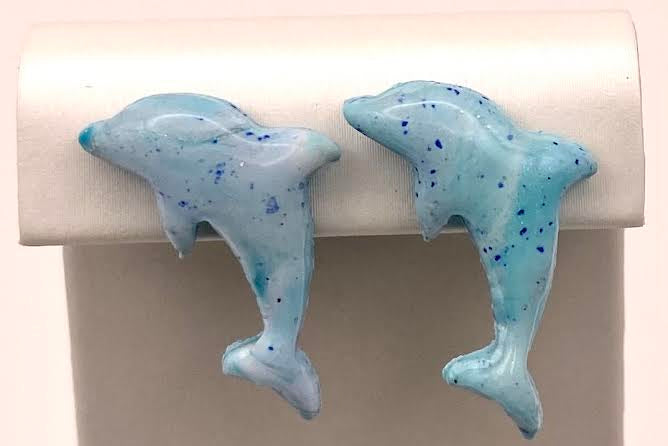 A pair of light blue dolphin-shaped earrings with dark blue speckles, displayed on a white jewelry stand against a light background.