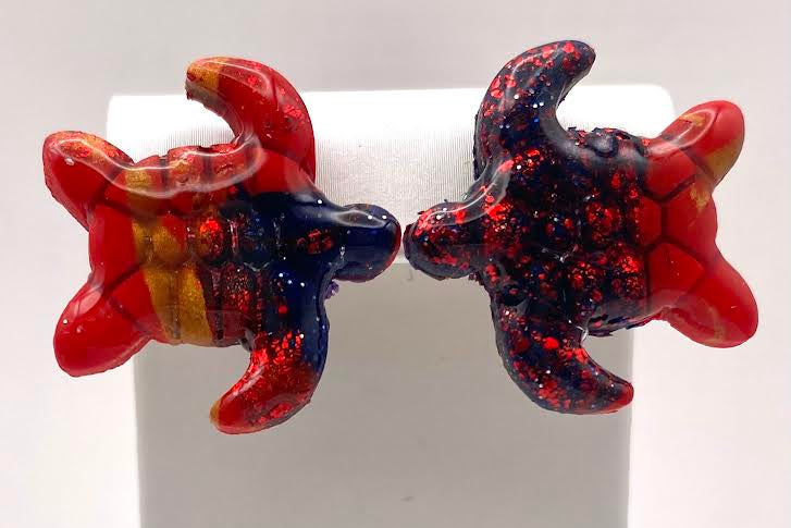A pair of handmade turtle earrings in red, navy blue, and gold, displayed on a white jewelry stand against a white background.