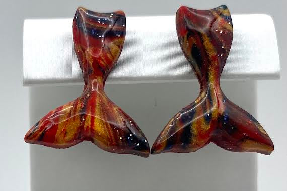 A pair of handmade glass earrings shaped like mermaid tails, featuring a marbled pattern of red, orange, yellow, and black, displayed on a white jewelry stand.