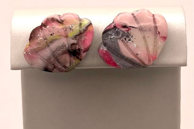 A pair of handmade heart-shaped stud earrings with a pink, white, and gray marbled pattern and small black speckles, displayed on a white stand against a light background.