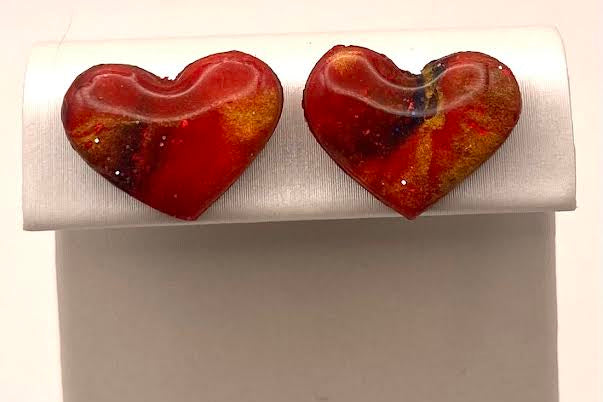 A pair of heart-shaped glass stud earrings with a marbled red, orange, and yellow pattern, displayed on a white jewelry stand.
