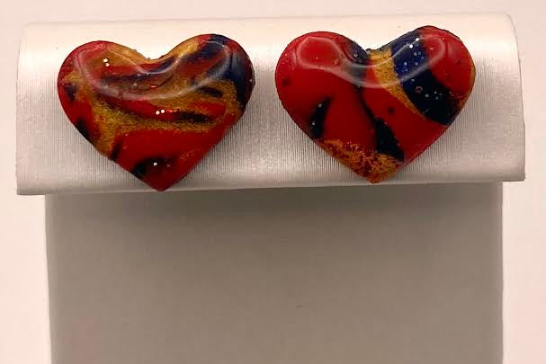 A pair of heart-shaped glass stud earrings with a marbled pattern of red, orange, yellow, and dark blue, displayed on a white jewelry stand.