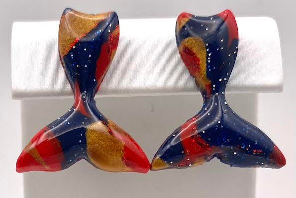 A pair of handmade mermaid tail stud earrings with a marbled pattern of navy blue, red, and gold, displayed on a white jewelry stand.