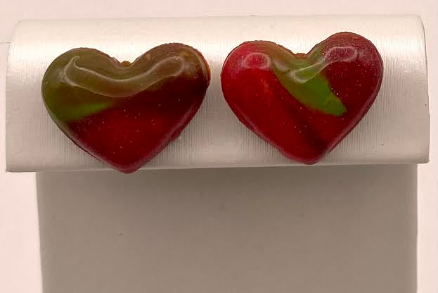 A pair of heart-shaped red and green glass stud earrings displayed on a white jewelry stand against a light background.