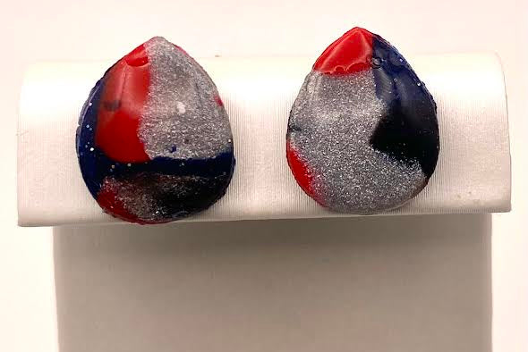 A pair of teardrop-shaped earrings with a marbled pattern of red, navy blue, and glittery silver, displayed on a white jewelry stand.