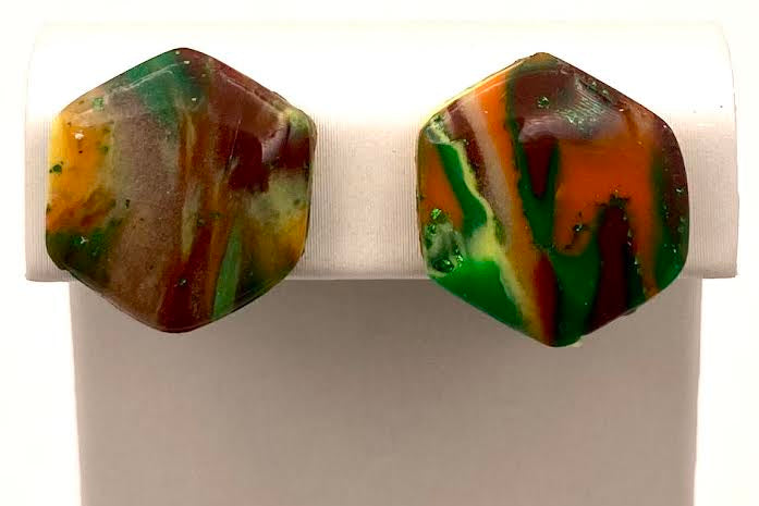 A pair of marbled hexagonal stud earrings with swirls of green, orange, brown, and yellow, displayed on a white rectangular stand.