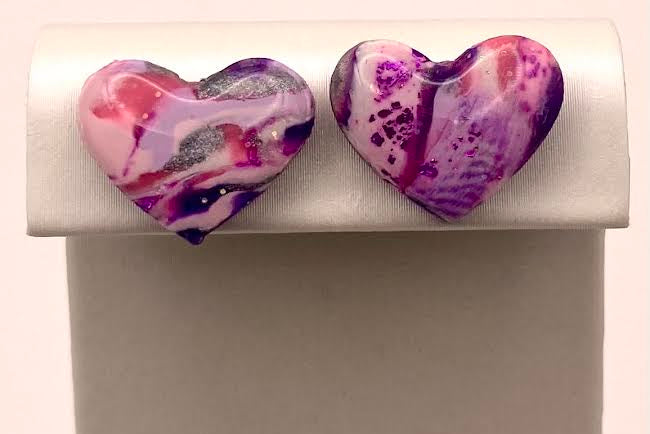 Two heart-shaped marbled resin jewelry charms in pink, purple, and white, displayed on a white jewelry stand.