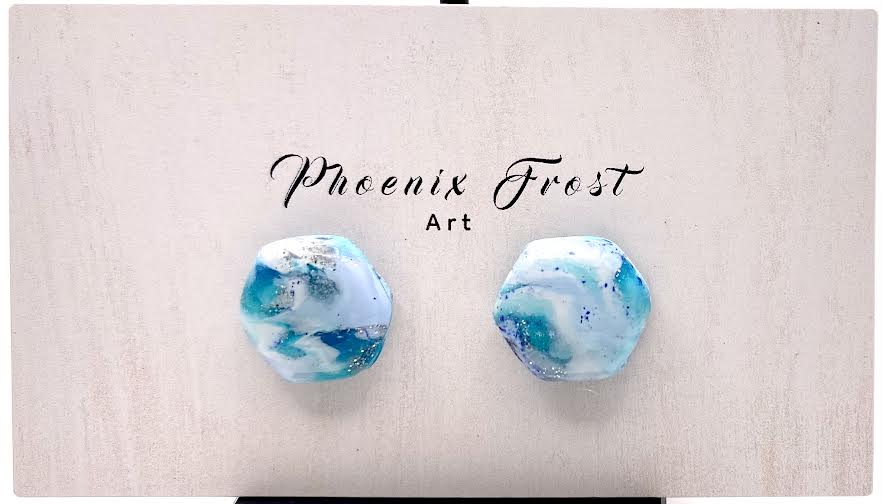 A pair of hexagon shaped earrings with a marbled blue, white, and light teal pattern, displayed on a light beige wooden card with the brand name 'Phoenix Frost Art' printed above them.